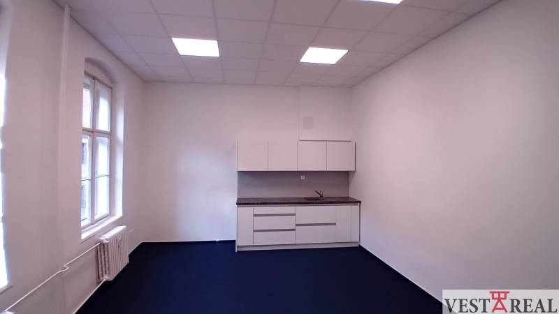 Kanceláře s kuchyňskou linkou a modrým kobercem, bílé stěny a velké okno.