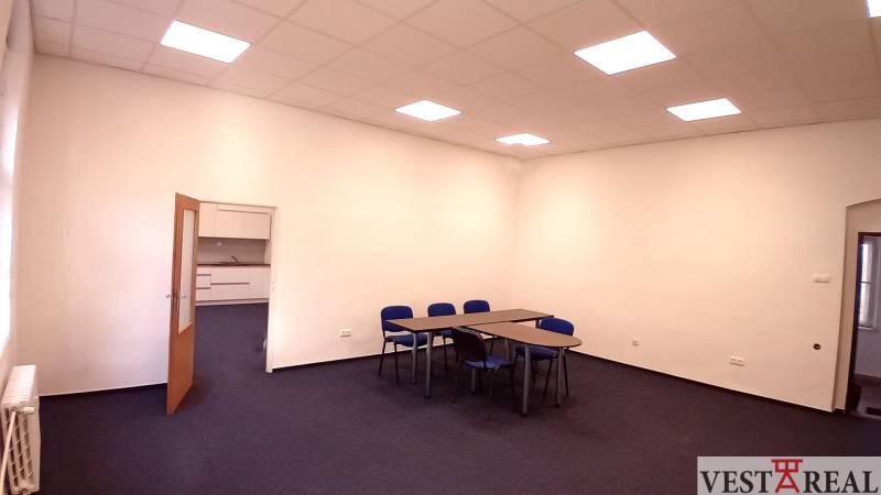 Kancelář s modrými židlemi a stoly, bílá stěna, otevřené dveře k malé kuchyňce.