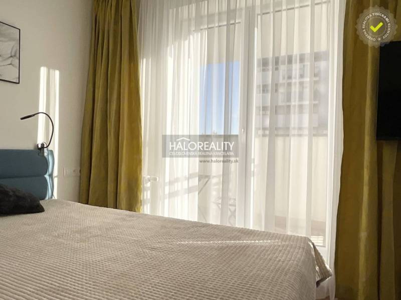 Světlá ložnice v jednopokojovém bytě s velkým oknem a závěsy.