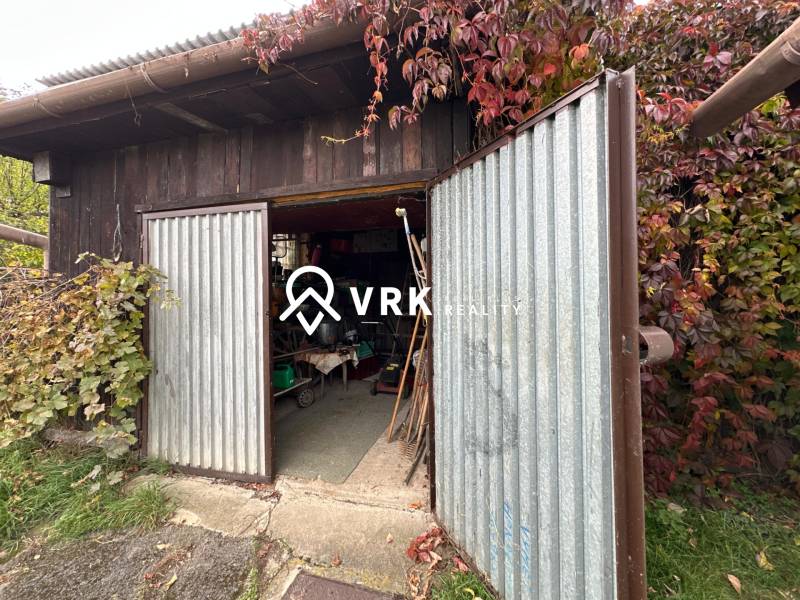 Otevřené kovové dveře garáže u rodinného domu v Kalši, obklopené bujnou vegetací.