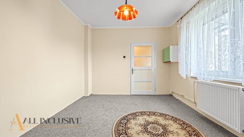 Světlý pokoj v rodinném domě s kobercem a oknem s krajkovou záclonou.