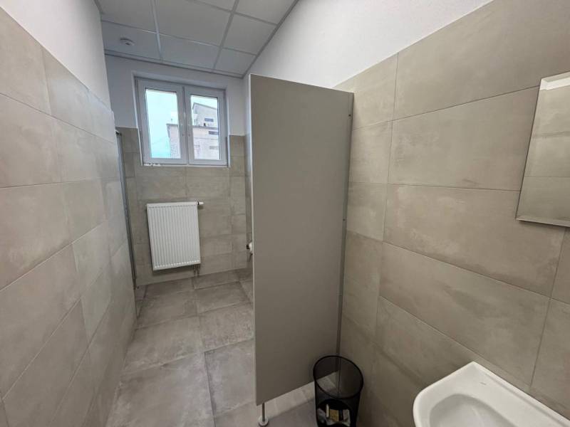 Kanceláře s šedým obkladem a toaletou, s malým oknem a žebrovým radiátorem.