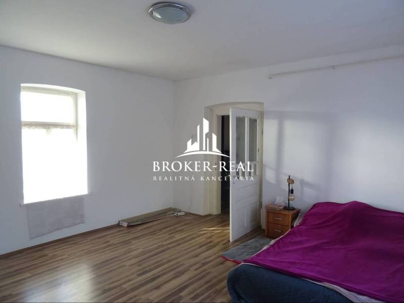 Pokoj s postelí, oknem, podlahou s dřevěným dekorem, nočním stolkem a otevřenými dveřmi.