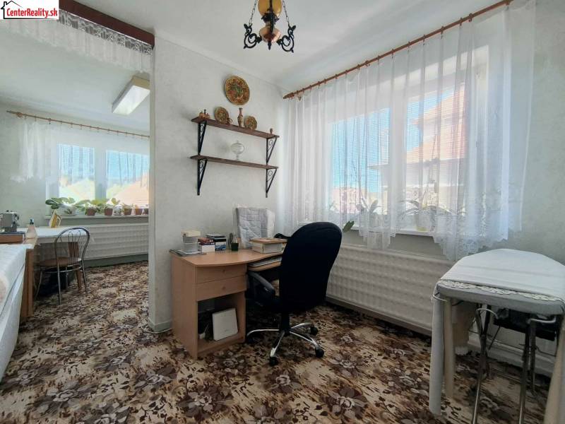 Pokoj v rodinném domě s psacím stolem, rostlinami a kobercem s květinovým vzorem.