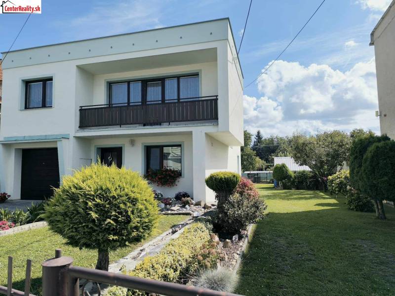 Rodinný dům v Partizánském obklopený zahradou s dřevinami a zeleným trávníkem.