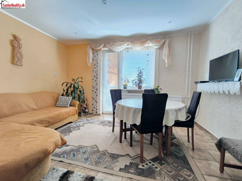 Obývací pokoj rodinného domu s jídelním stolem, gaučem a televizí na polici.