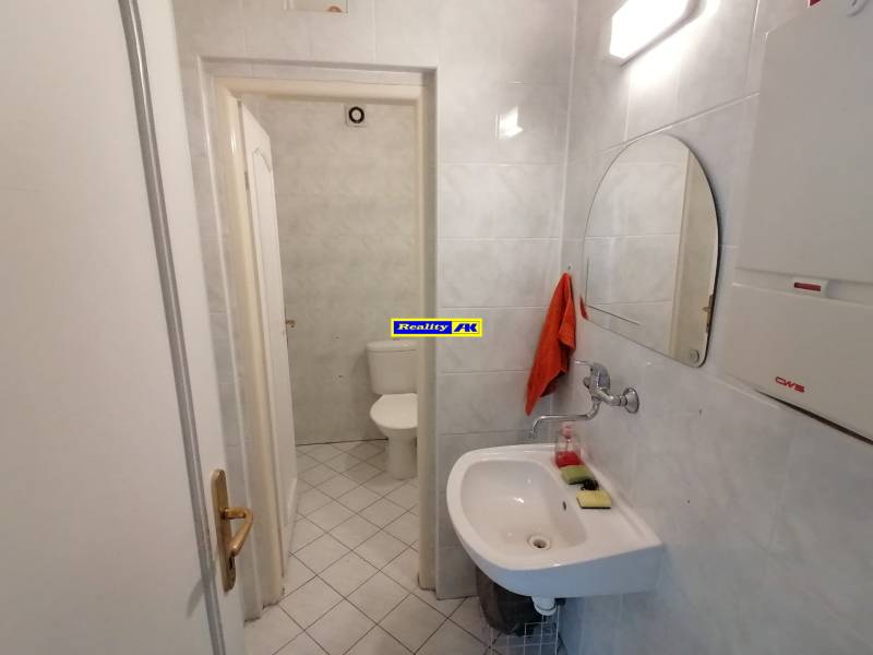 Koupelna v kancelářích se zrcadlem, umyvadlem a toaletou na bílých obkladech.