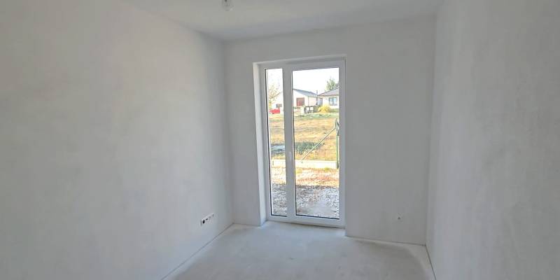 Bílá místnost v rodinném domě s velkým oknem a výhledem na zahradu.