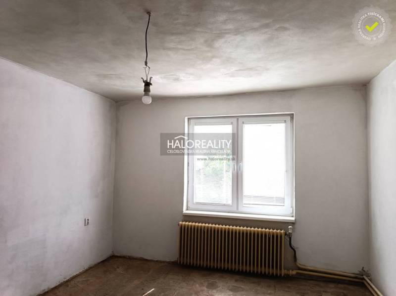 Prázdná místnost v rodinném domě s bílými stěnami, radiátorem a oknem.