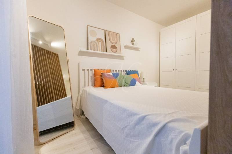 Ložnice v jednopokojovém bytě s postelí, barevnými polštáři, zrcadlem a velkou skříní.