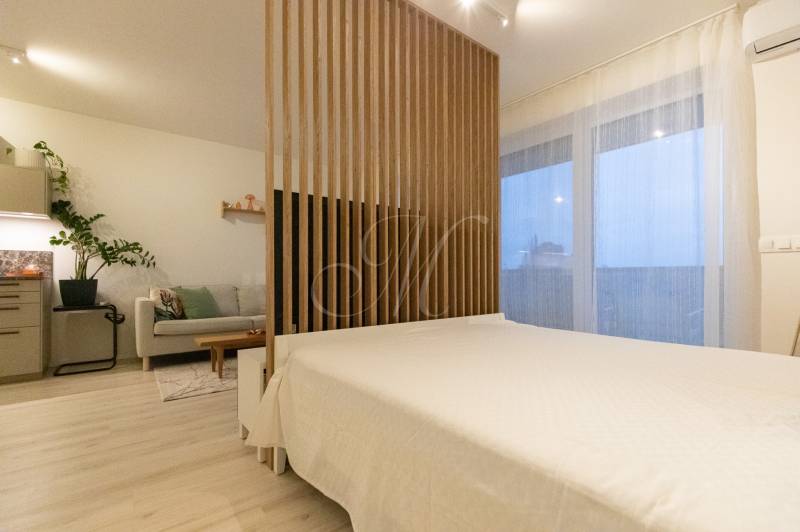 Kuchyně se světlým nábytkem, pohovkou a lůžkem v 1-pokojovém bytě, podlaha s dřevěným dekorem.