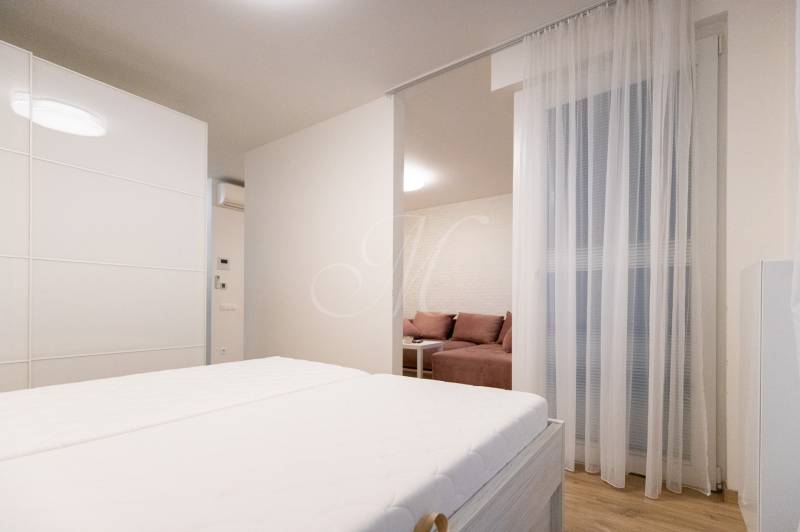 Dvoupokojový byt s postelí, podlahou s dřevěným dekorem a rozdělenou obývací částí.