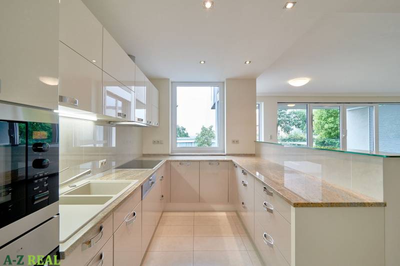 Kuchyně v 3-pokojovém bytě s velkým oknem, bílými skříňkami a lesklou pracovní deskou.