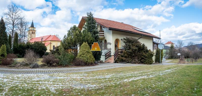 Vila na ulici Andreje Sládkoviče v Žarnovici, obklopená zelení, s kostelem v pozadí.