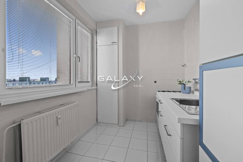 Kuchyně v 1-pokojovém bytě s bílým nábytkem a kachličkami, s výhledem přes žaluzie.