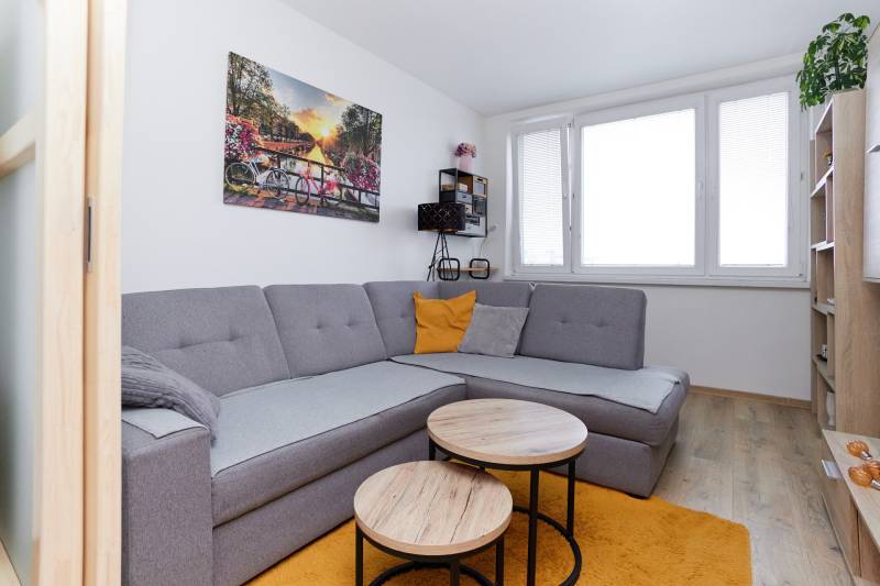 Obývací pokoj dvoupokojového bytu s rohovou sedačkou, podlahou s dřevěným dekorem a oknem.