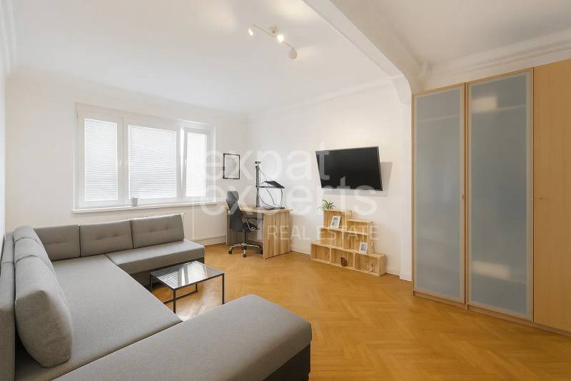 Obývací pokoj ve 3-pokojovém bytě s rohovou sedačkou, pracovním stolem a podlahou s dřevěným dekorem.