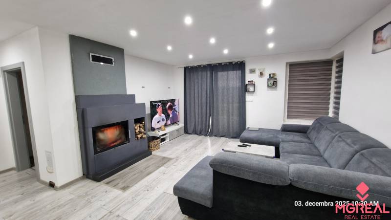 Obývací pokoj v rodinném domě s krbem, šedou sedačkou a televizorem, podlaha s dřevěným dekorem.