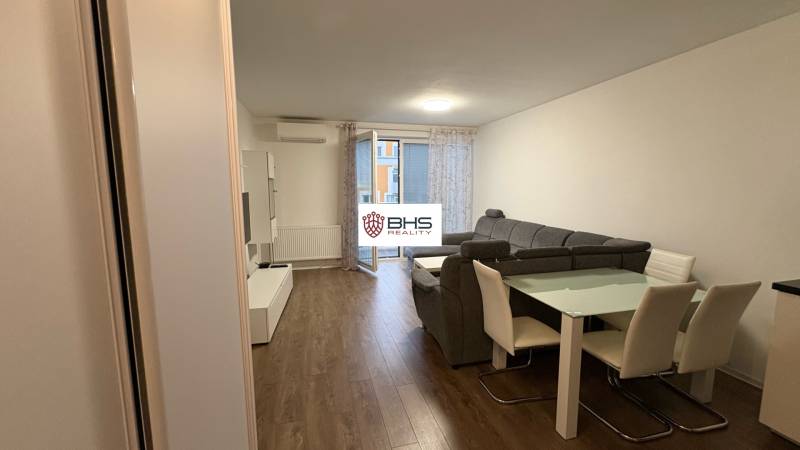 Obývací pokoj v jednopokojovém bytě s podlahou s dřevěným dekorem, šedou sedačkou a jídelním stolem.