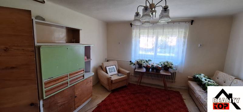 Obývací pokoj v rodinném domě s křeslem, starší skříní a stolem s květinami.