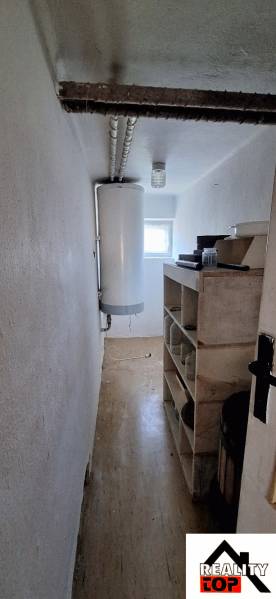 Úzký interiér rodinného domu s bojlerem a poličkami, světlé stěny, podlaha s dřevěným dekorem.