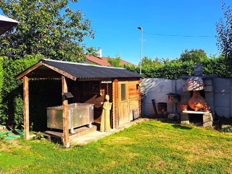 Zahrada rodinného domu v Nedede s dřevěným domkem a venkovním krbem.