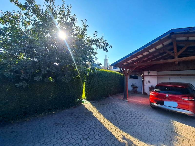 Rodinný dům v Nedede s červeným autem před garáží, slunečný den a strom.