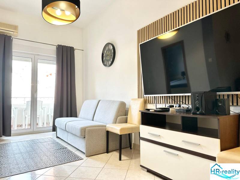 Obývací pokoj s pohovkou a televizorem v rekreačním apartmánu, světlá dlažba, balkónové dveře.