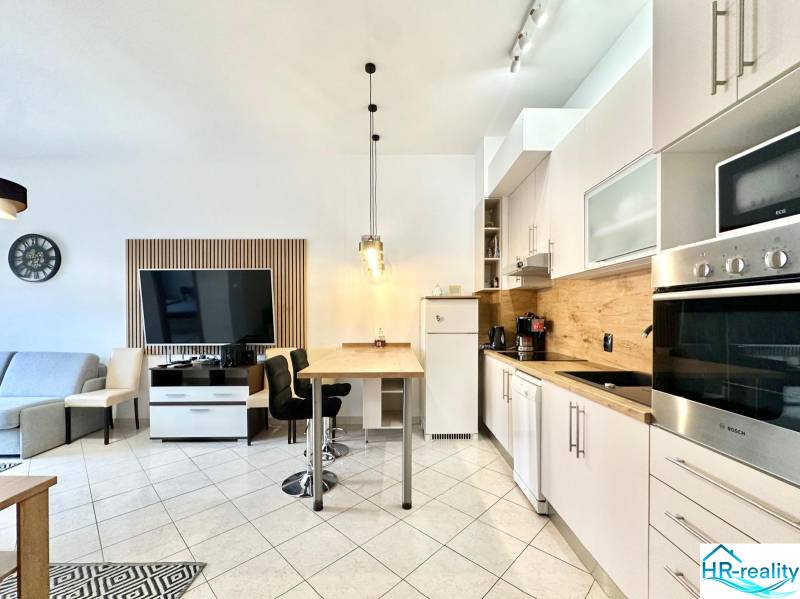 Interiér rekreačního apartmánu s kuchyňskou linkou, jídelním stolem a televizí na stěně.