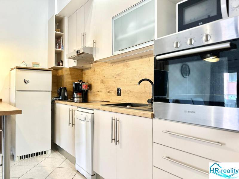 Kuchyně v rekreačním apartmánu s bílým nábytkem a dřevěným dekorem na stěně.