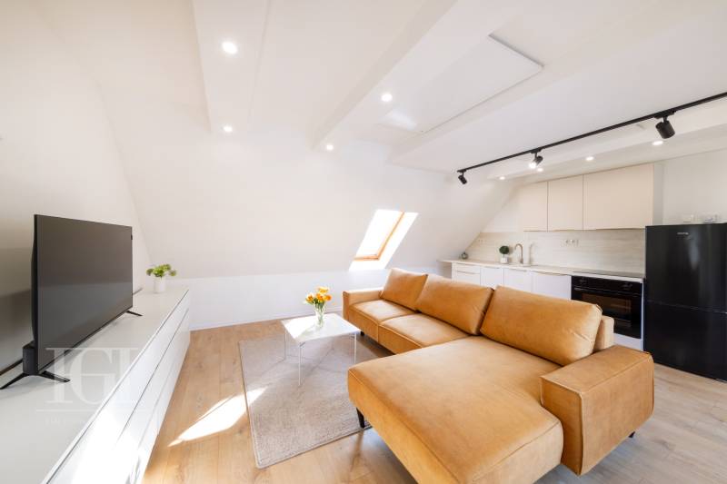 Obývací pokoj s kuchyní ve 2pokojovém bytě: velké okno, hnědá sedačka, podlaha s dřevěným dekorem.