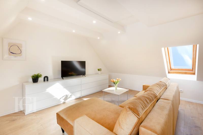 Obývací pokoj v dvoupokojovém bytě s pohovkou, televizí a střešním oknem, bílá komoda.