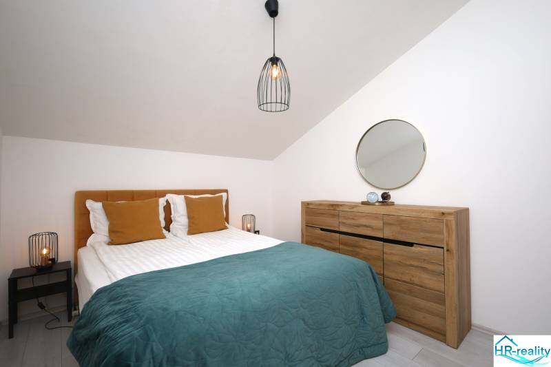 Ložnice v rekreačním apartmánu s postelí, dřevěnou komodou, zrcadlem a lampou.