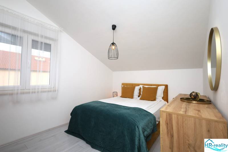 Ložnice v rekreačním apartmánu s manželskou postelí, oknem a komodou pod šikmou střechou.