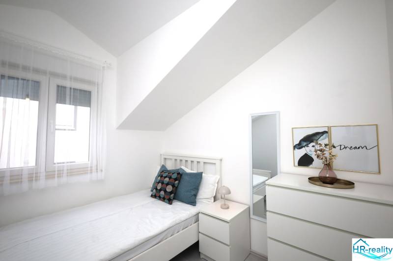 Světlá ložnice v rekreačním apartmánu s postelí, nočním stolkem a komodou.