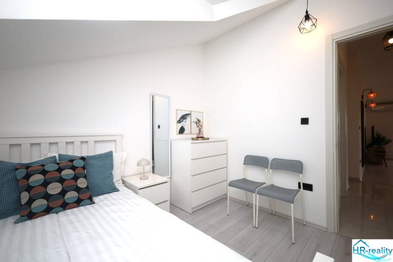 Rekreační apartmán se světlou ložnicí, postelí, komodou a polštáři, podlaha s dřevěným dekorem.