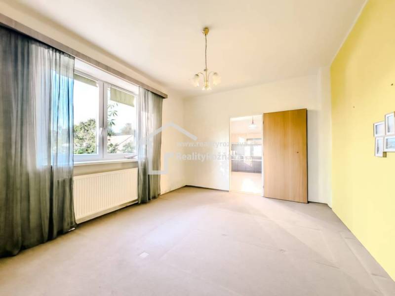 Pokoj s velkým oknem, radiátorem a průchodem v rodinném domě.