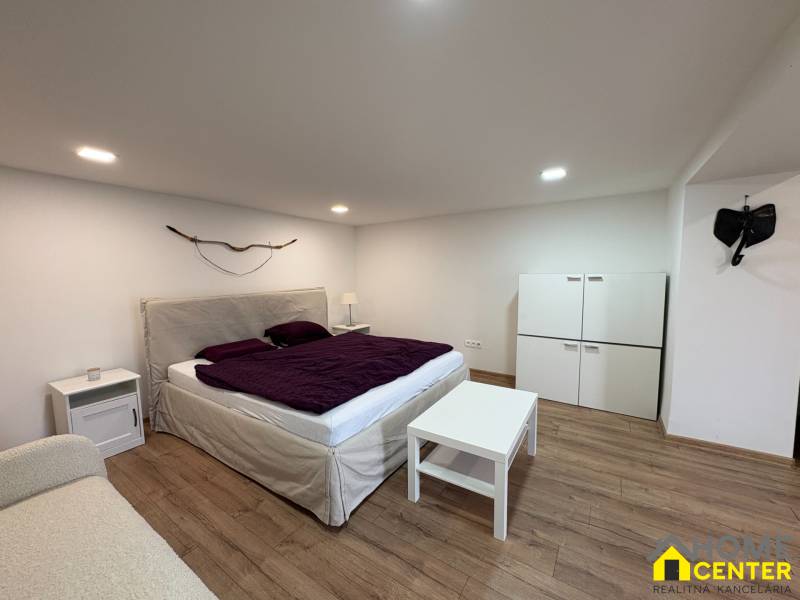 Ložnice v jednopokojovém bytě s podlahou s dřevěným dekorem a postelí s fialovým přehozem.