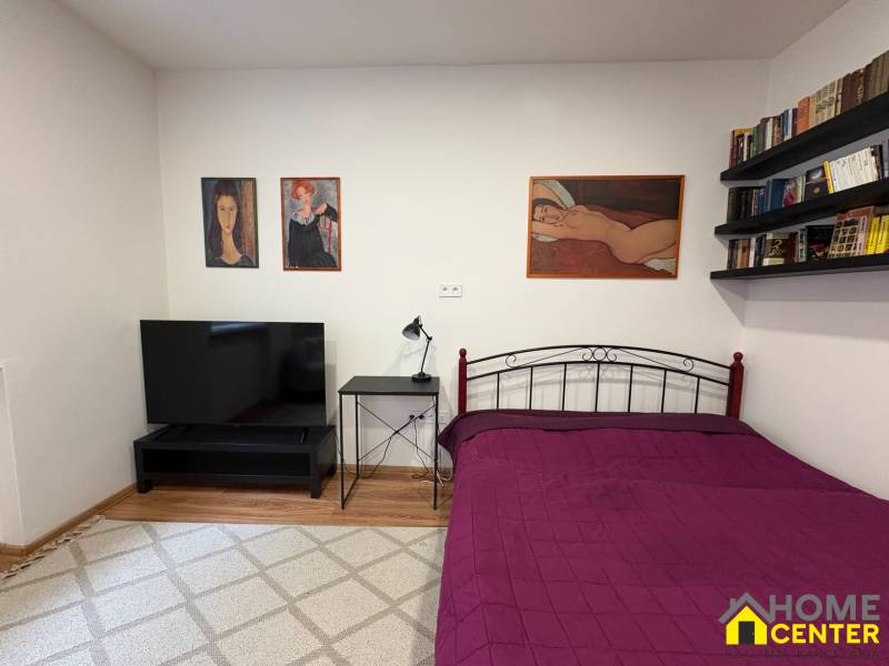 Ložnice v jednopokojovém bytě s televizorem a obrazy na strohých stěnách.