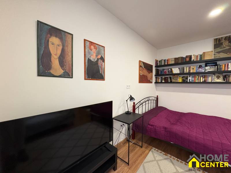 Jednopokojový byt s postelí, policemi s knihami a televizorem, podlaha s dřevěným dekorem.