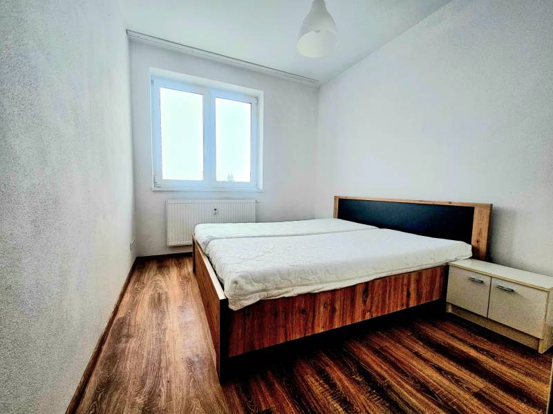 Ložnice v dvoupokojovém bytě s podlahou s dřevěným dekorem, postelí a oknem.