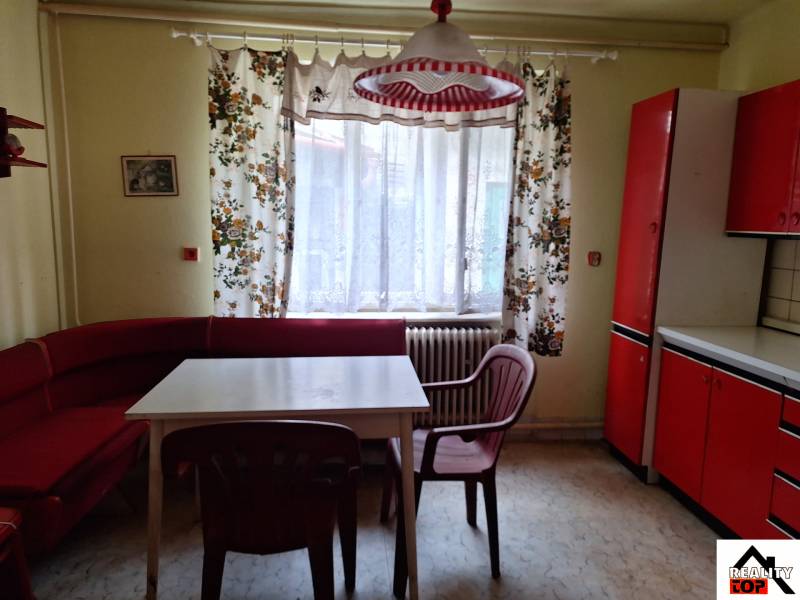 Kuchyně v rodinném domě s červeným nábytkem, barevnými závěsy a plastovými židlemi.