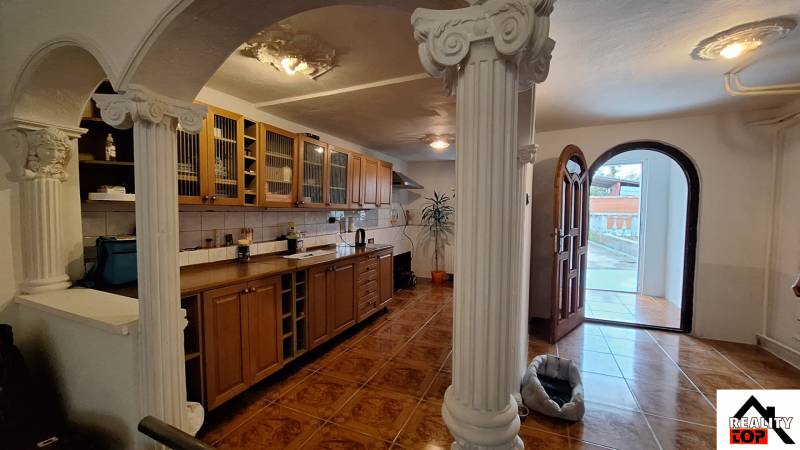 Interiér rodinného domu s dřevěnou kuchyňskou linkou, sloupy a podlahou s dřevěným dekorem.