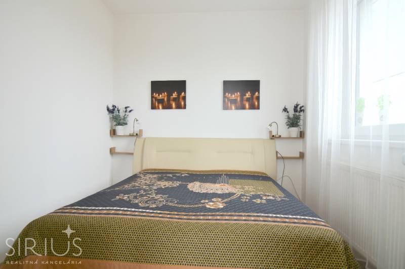 Ložnice v dvoupokojovém bytě se světlou postelí a dekorativními obrázky na stěně.