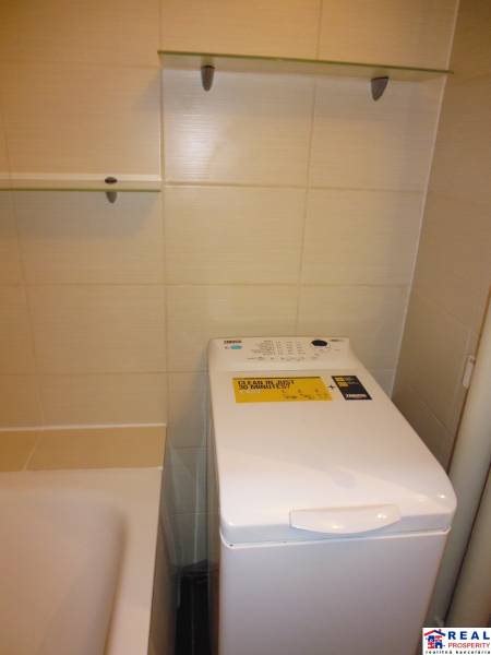 Koupelna v 2-pokojovém bytě s pračkou vedle vany a policemi.