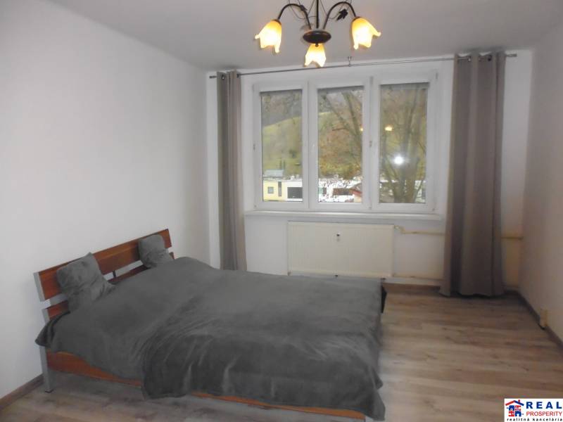 Ložnice v dvoupokojovém bytě s postelí a podlahou s dřevěným dekorem, šedé závěsy.