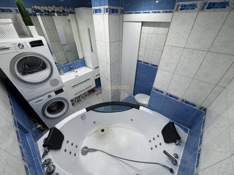 Koupelna v 3-pokojovém bytě s rohovou vanou, pračkou, sušičkou a umyvadlem.