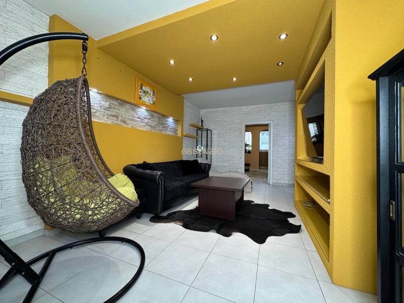 Obývací pokoj v 3pokojovém bytě s houpacím křeslem, černou pohovkou a žlutými stěnami.