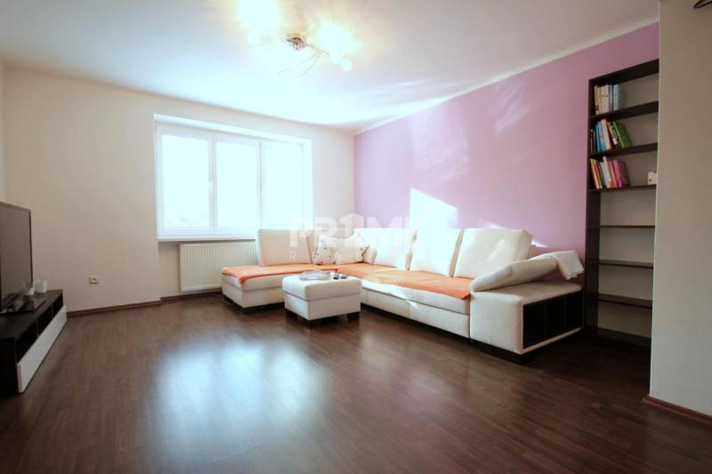 Obývací pokoj dvoupokojového bytu s rohovou sedačkou, knihovnou a podlahou s dřevěným dekorem.