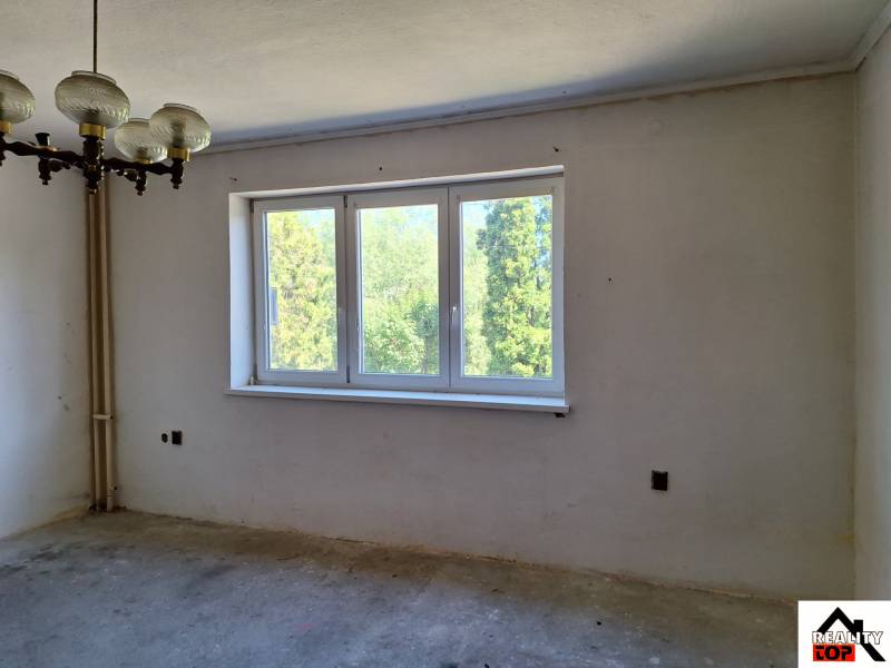 Pokoj v rodinném domě s velkým oknem, původním lustrem a výhledem na zeleň.